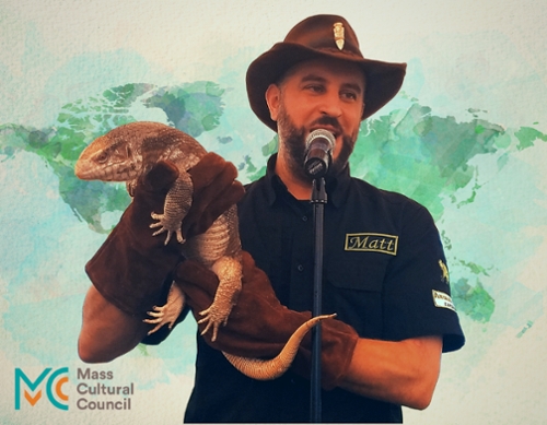 Image of a man in a brown ranger hat wearing large brown gloves and holding a large lizard.