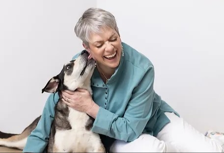 A smiling woman with short grey gair and a white and black dog licking her neck.